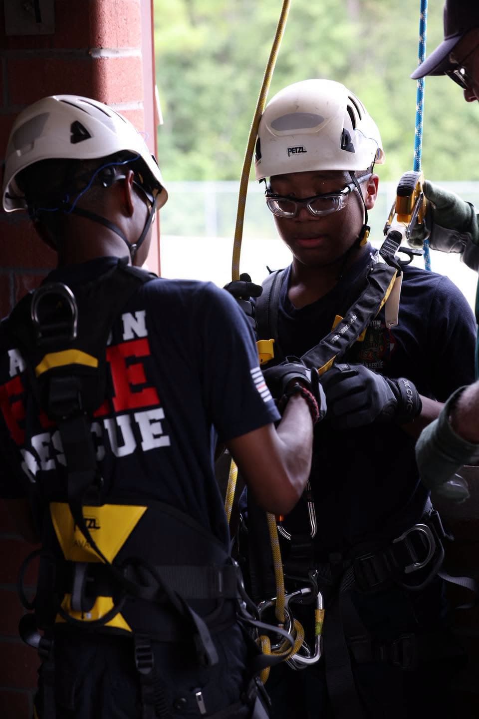 Two helmeted Gaston cadets wearing harnesses and handling rope equipment beside a window